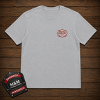 A light heather gray crewneck t-shirt with a small red embroidered 'M&M Custom Company' logo on the left chest, displayed flat on a dark wood plank background with an M&M patch graphic in the bottom-left corner.
