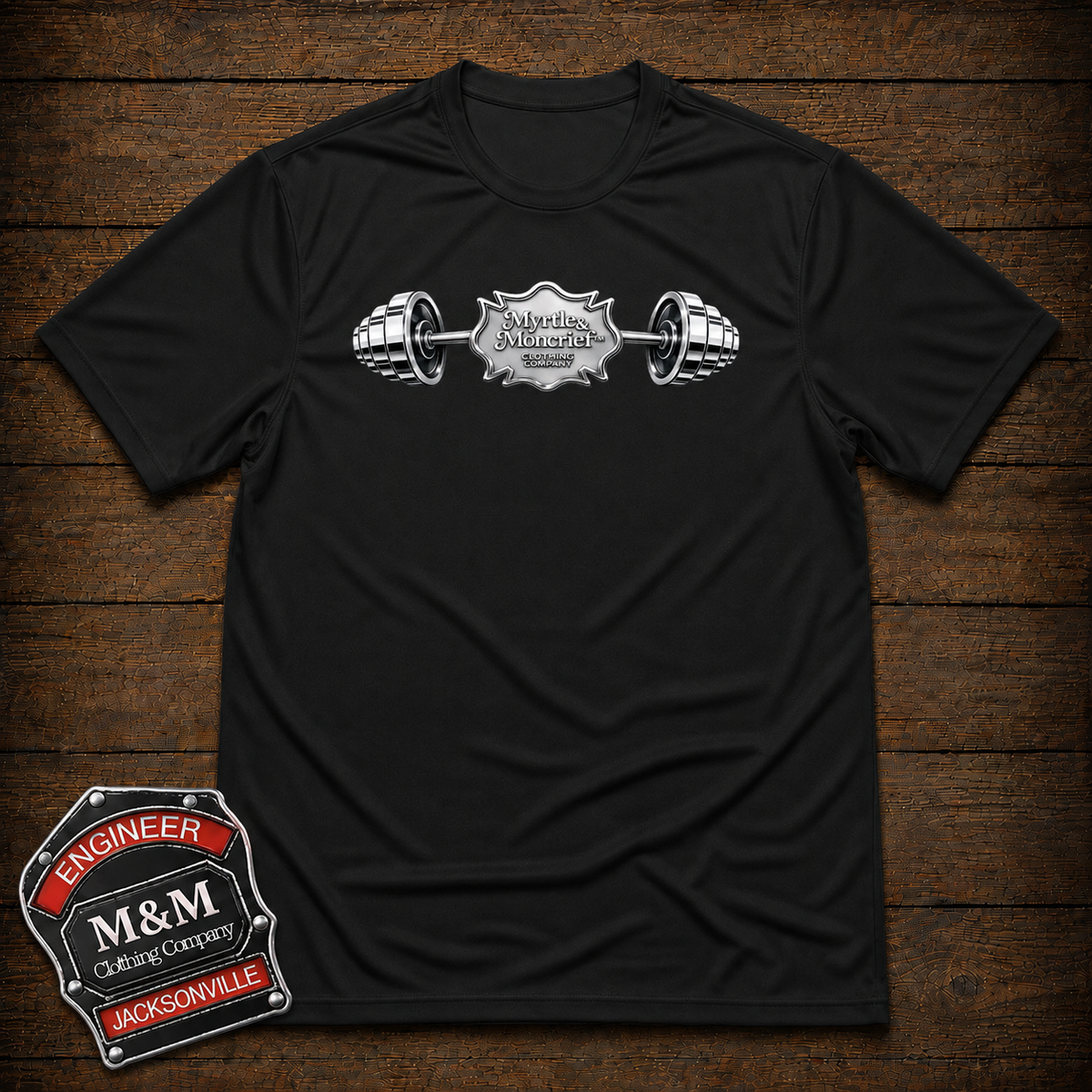A black short-sleeve crewneck t-shirt with a silver and white barbell graphic across the chest featuring the text 'M&M's Motorcycle Engineering Corporation'. The shirt is displayed flat on a dark brown wooden plank background, with an 'Engineer M&M's' badge overlay in the bottom-left corner.