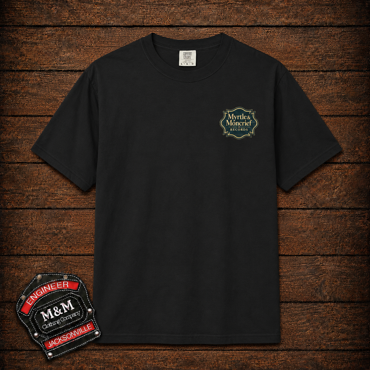 A black short-sleeve crewneck t-shirt with a small 'Mortley Monster' graphic on the left chest, displayed flat against a dark wood plank background. A decorative 'Engineer M&M Escapable' patch is shown in the bottom-left corner.