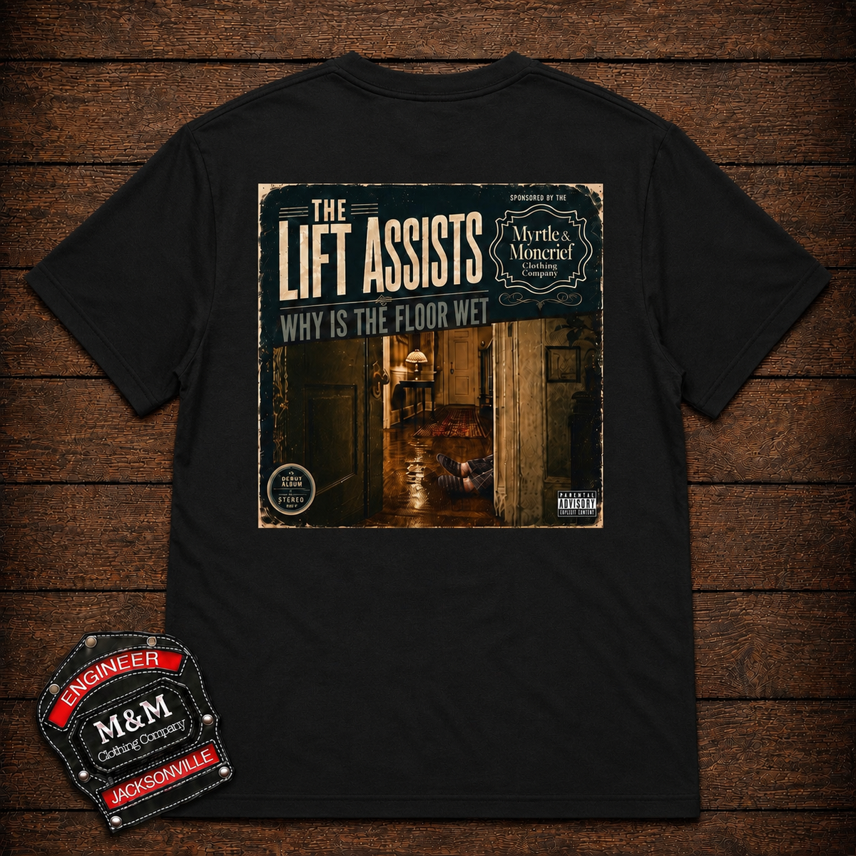 A black short-sleeve graphic t-shirt laid flat on a dark wood plank background. The shirt features a large rectangular front print with the text 'THE LIFT ASSISTS' and 'WHY IS THE FLOOR WET' above an image of a dimly lit hallway.