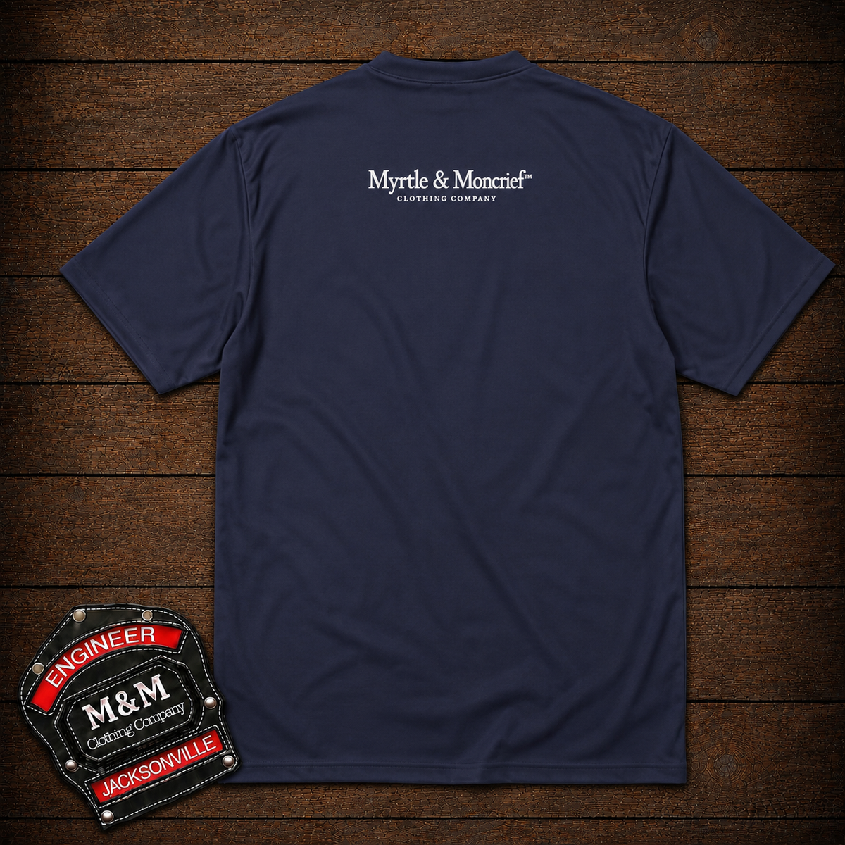 A navy blue short-sleeve t-shirt shown from the back, laid flat on a dark wood plank background. The shirt features a white printed logo for 'Myrtle & Monrief' on the upper back.