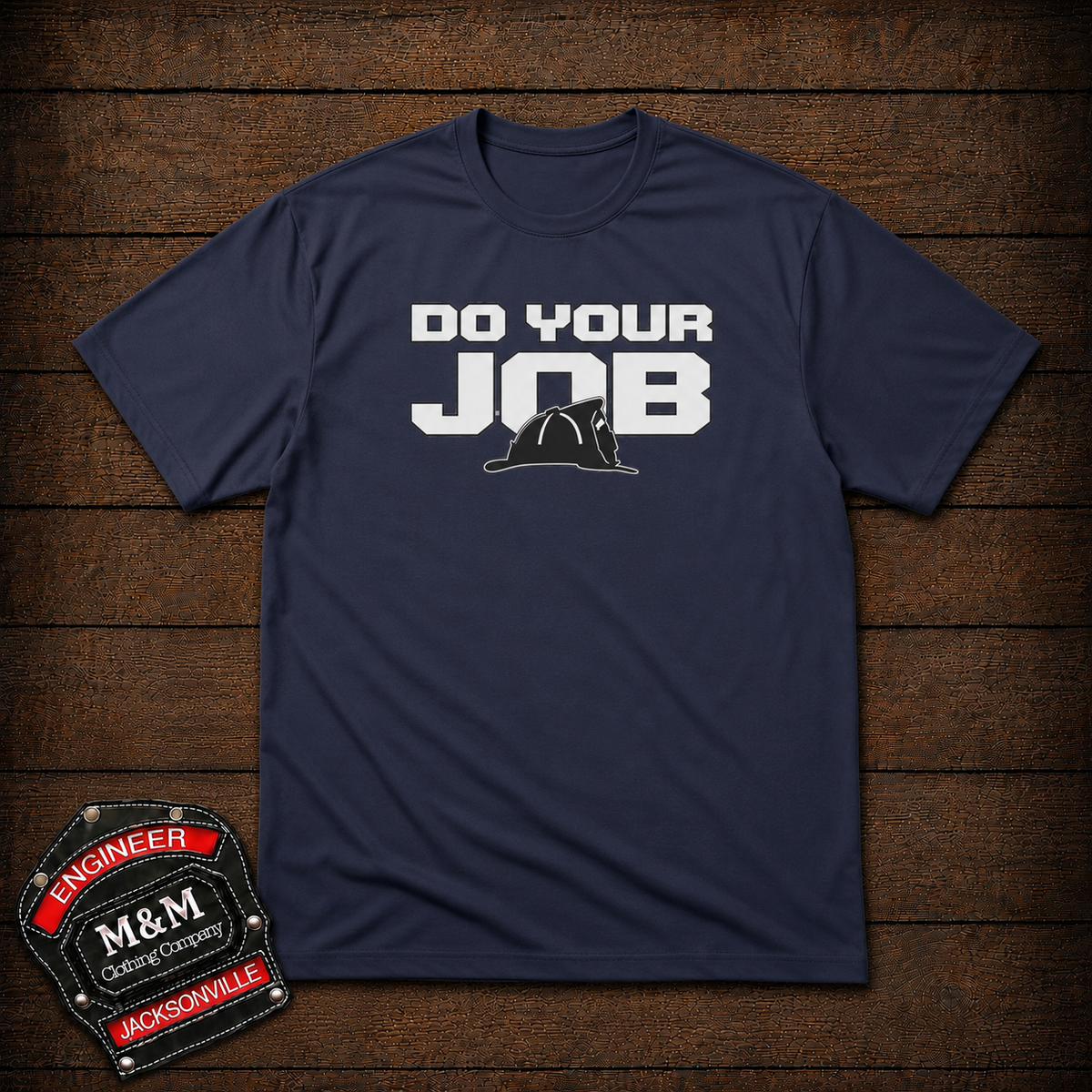 A navy blue short-sleeve t-shirt with a graphic print that reads 'DO YOUR JOB' in white, featuring a black firefighter helmet silhouette. The shirt is displayed flat on a dark wood plank background, with an 'M&M Engineer' badge overlay in the bottom-left corner.
