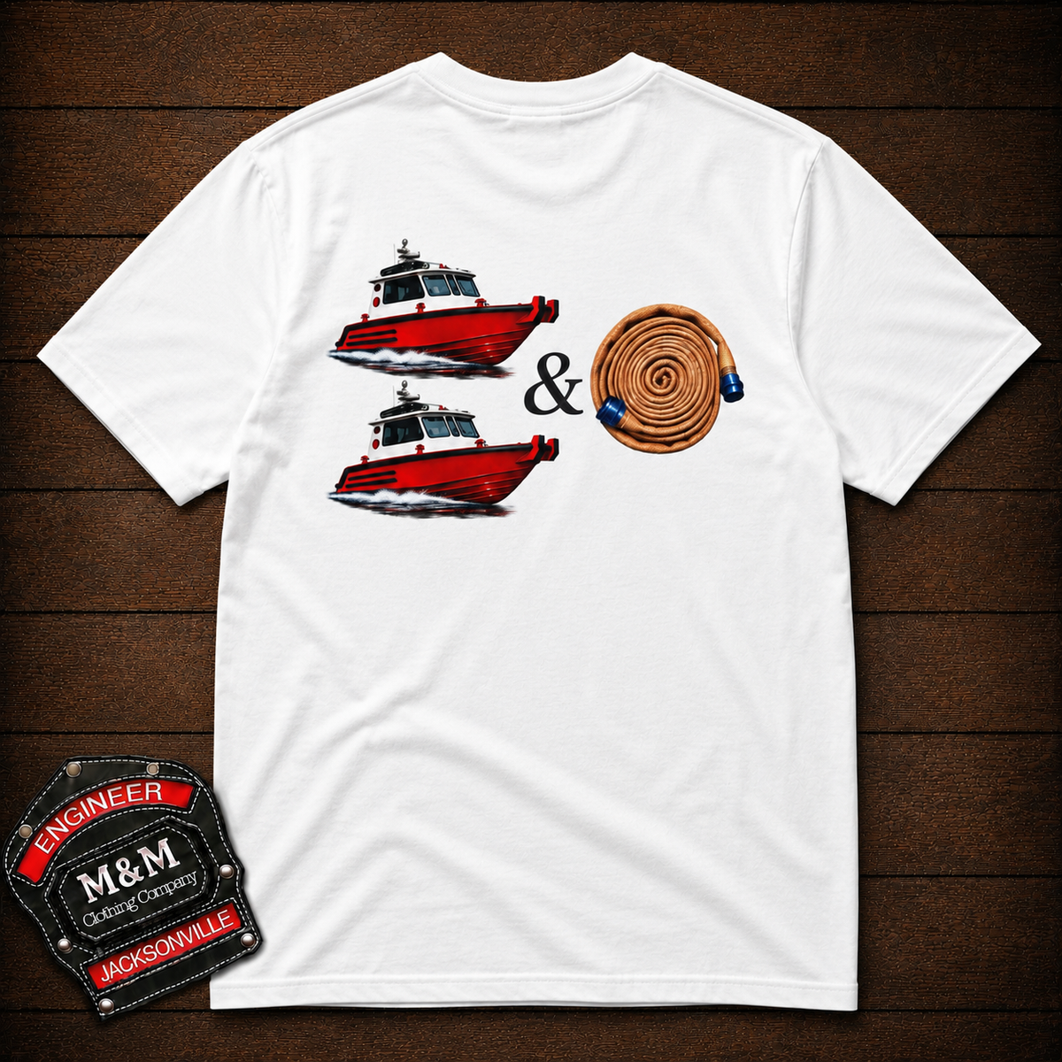 A white short-sleeve t-shirt laid flat on a dark wood background, featuring a graphic print of two red and white boats, an ampersand symbol, and a coiled tan rope. An embroidered black and red 'Engineer M&M Jacksonville' patch is visible in the bottom-left corner.