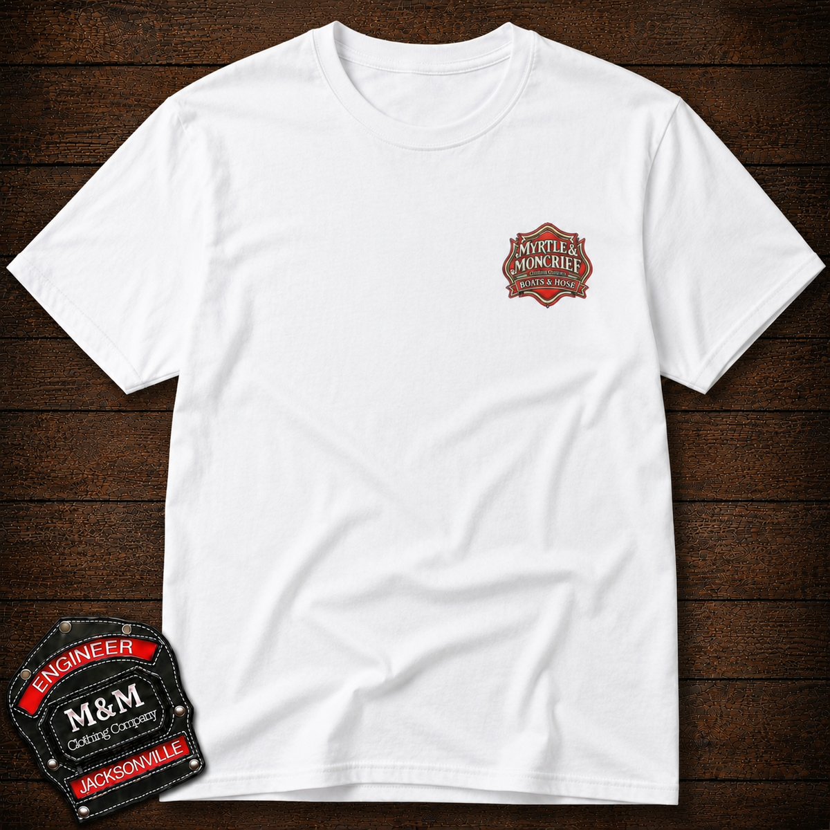 A white short-sleeve crewneck t-shirt laid flat on a dark wood background, featuring a small red and black 'Minty's Moncrief' logo on the left chest. A larger version of the 'M&M Engineer' logo patch is shown in the bottom-left corner as an overlay.