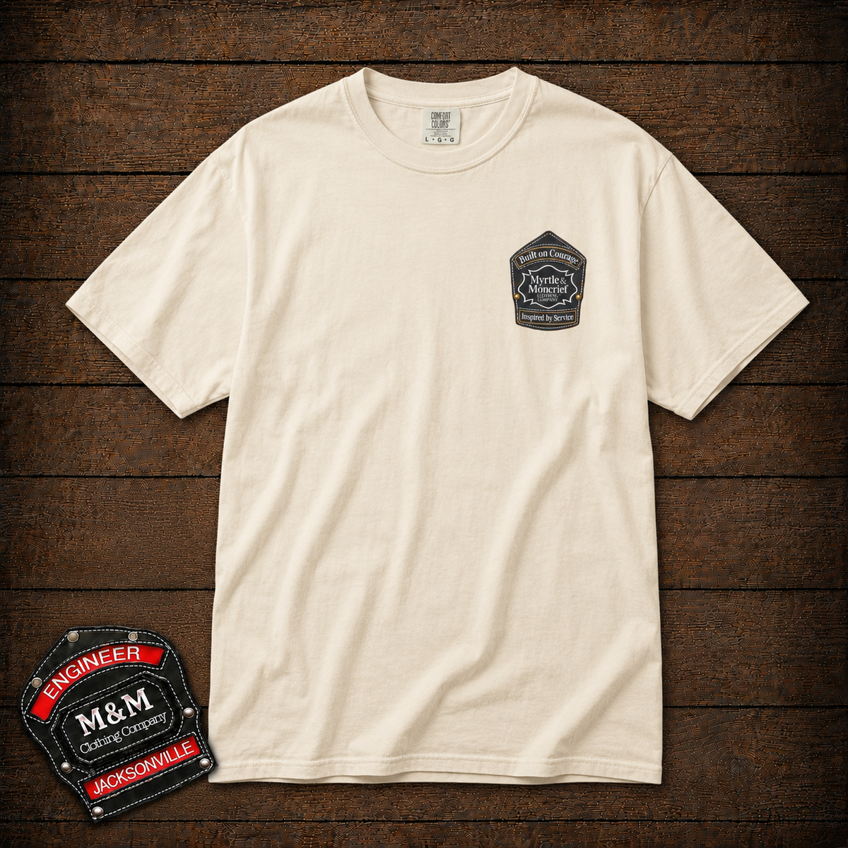 An off-white crewneck t-shirt with a small black and red 'M&M' Engineer patch printed on the left chest, displayed flat on a dark wood plank background with a matching patch graphic overlay in the bottom-left corner.