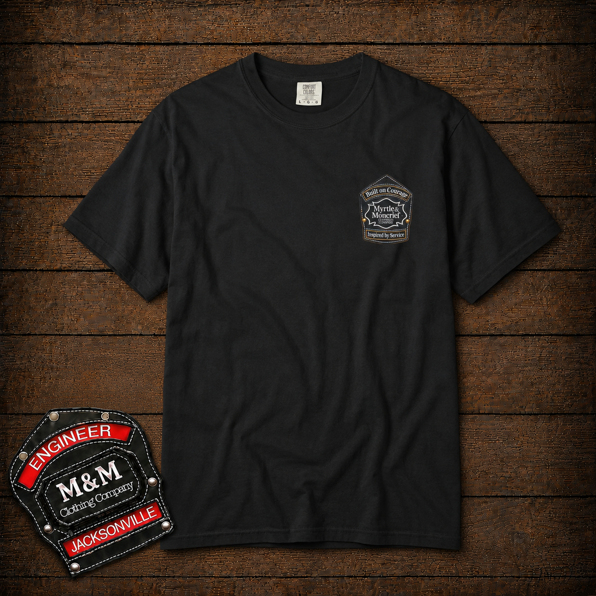 A black short-sleeve t-shirt with a small embroidered M&M Engineering Company patch on the left chest, displayed flat on a dark wood plank background. A larger version of the patch is shown as an overlay in the bottom-left corner.