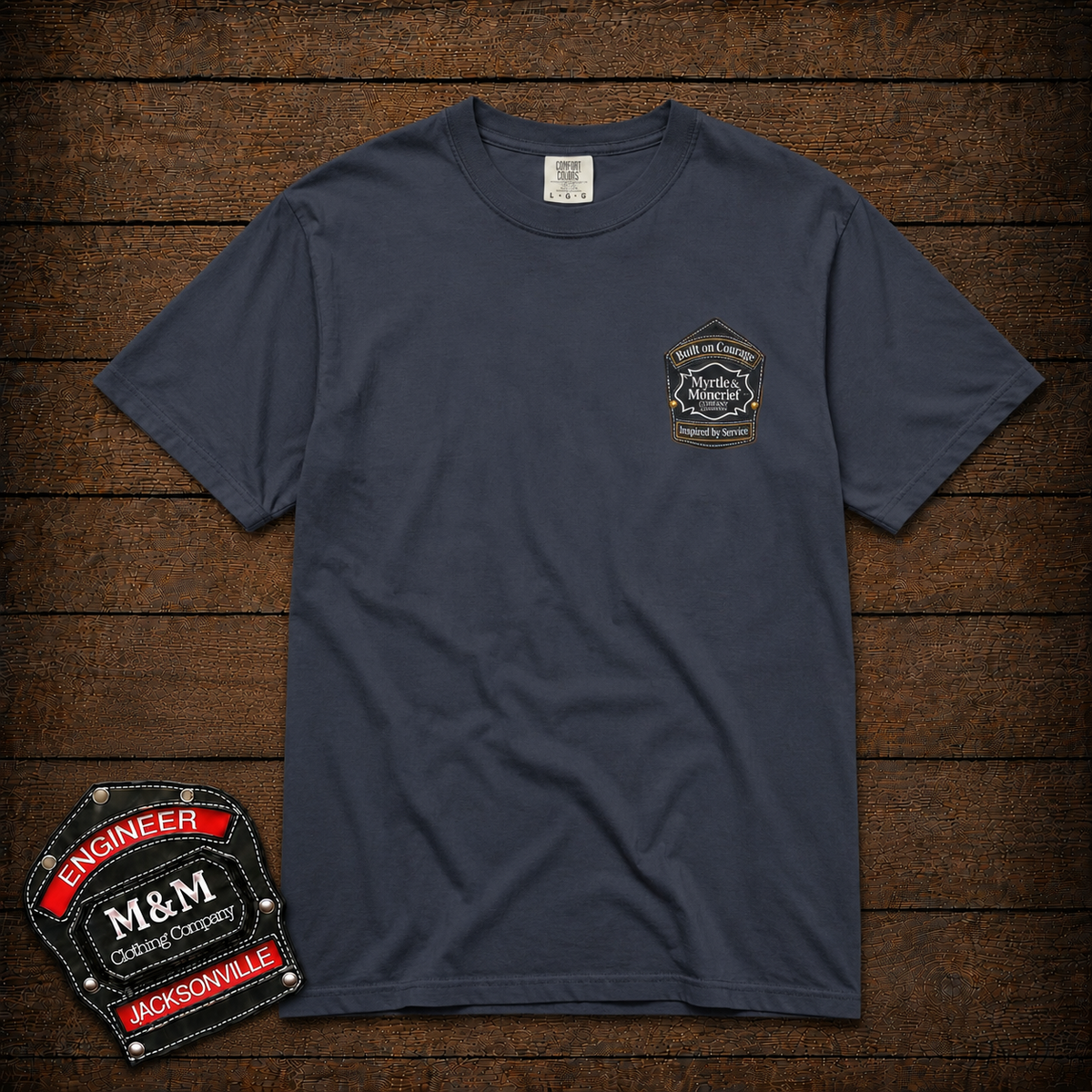 A navy blue short-sleeve t-shirt with a small embroidered patch on the left chest, displayed flat on a dark wood plank background. A larger embroidered patch is shown in the bottom-left corner as a graphic overlay.