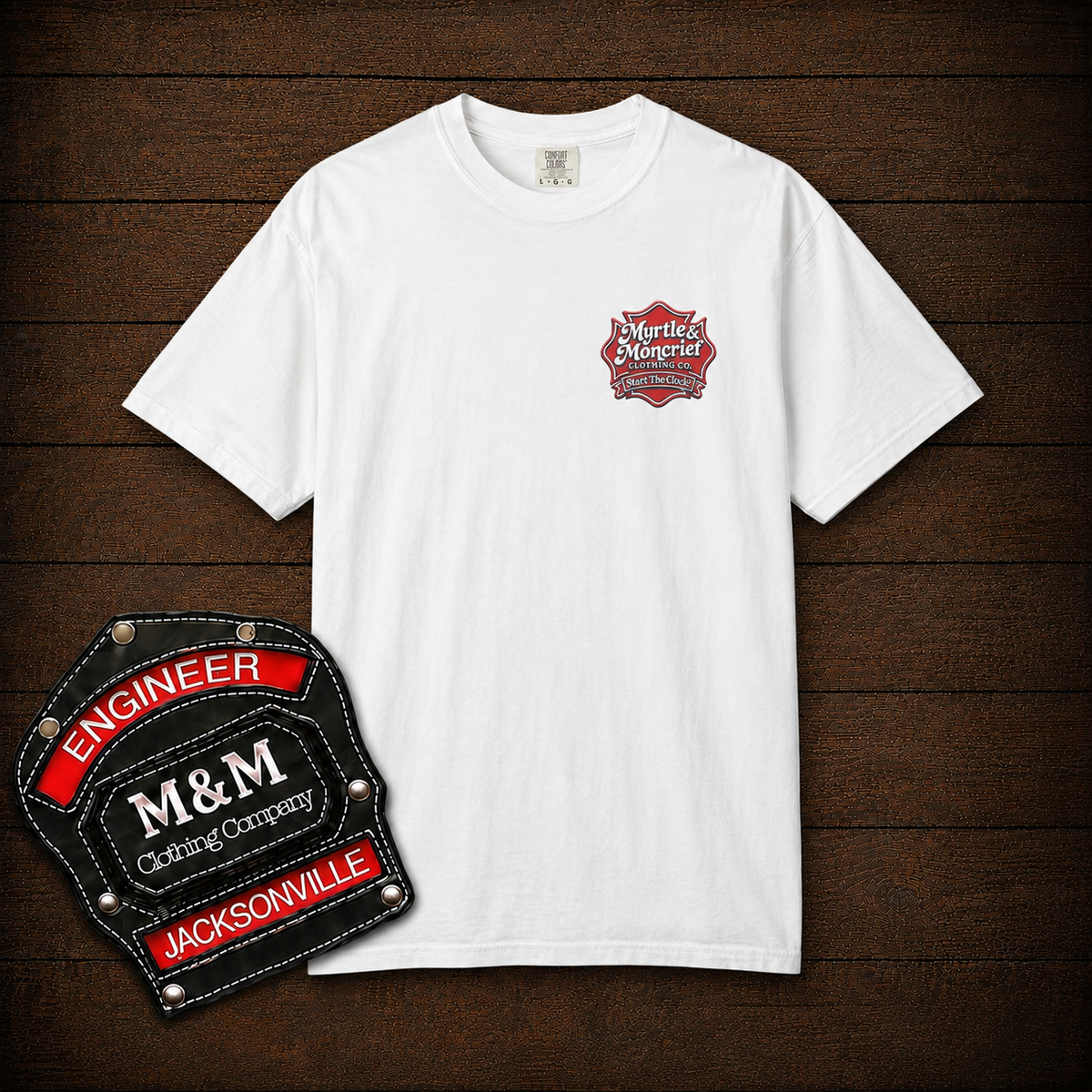 A white short-sleeve crewneck t-shirt with a small red and black 'M&M's Engineer' logo on the left chest, displayed flat on a dark wood background with a graphic badge overlay in the bottom left corner.