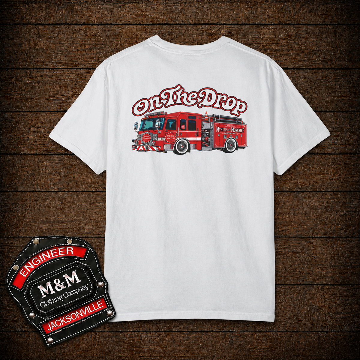 A white short-sleeve t-shirt with a graphic of a red fire truck and the text 'On The Drop' on the front, displayed flat on a dark wood background. A black and red 'Engineer M&M Racing Company' patch is shown in the bottom-left corner.
