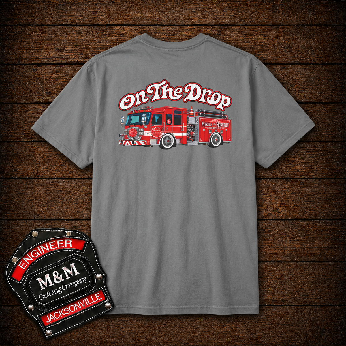 A gray short-sleeve t-shirt with a red fire truck graphic and the words 'On The Drop' on the back, displayed on a dark wood plank background. A black and red 'Engineer M&M Outfiting Company' patch is shown in the bottom-left corner.