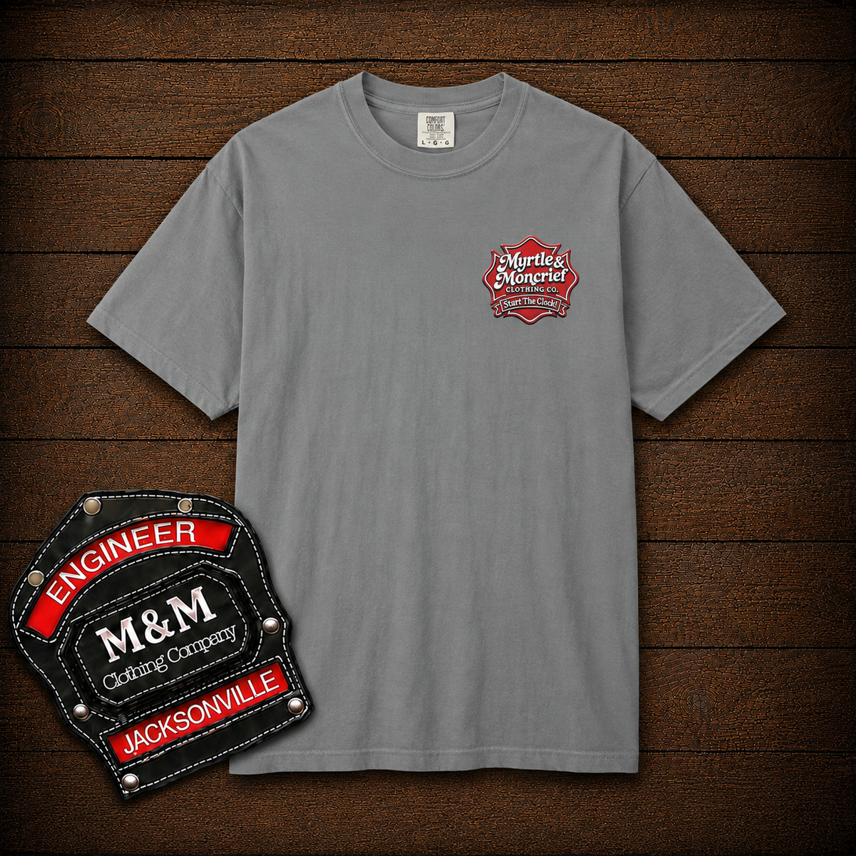 A gray short-sleeve crewneck t-shirt with a small red and white 'Mighty's Mangeriel' chest graphic, displayed on a dark wood plank background alongside a large embroidered patch graphic.
