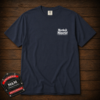 A navy blue short-sleeve t-shirt with a white graphic print on the left chest reading 'Myrtle & Moncrief' and 'BORN IN JACKSONVILLE', displayed flat on a dark wood plank background. A decorative embroidered patch for 'M&M Engineers Jacksonville' is placed in the bottom-left corner.
