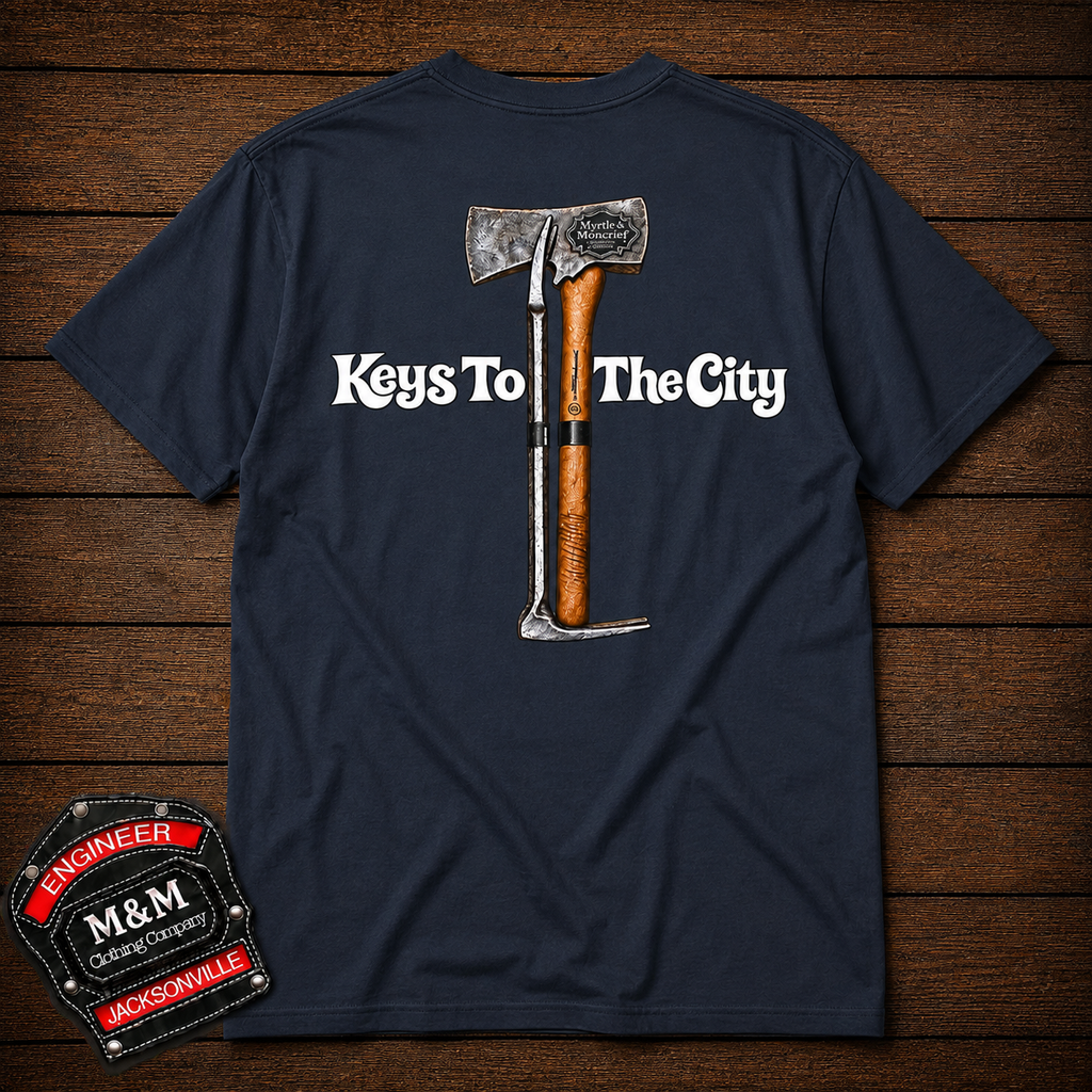 A navy blue t-shirt laid flat on a dark wood background, featuring a graphic of a tomahawk with the text 'Keys To The City' in white. An M&M Engineers Jacksonville patch is shown in the bottom-left corner.