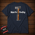 A navy blue t-shirt laid flat on a dark wood background, featuring a graphic of a tomahawk with the text 'Keys To The City' in white. An M&M Engineers Jacksonville patch is shown in the bottom-left corner.