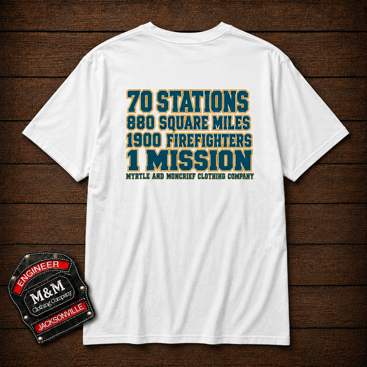 A white short-sleeve t-shirt with a vintage-style blue and yellow text graphic on the front, displayed flat on a dark wood background. A 'M&M Engineer Jacksonville' patch is shown in the bottom-left corner.