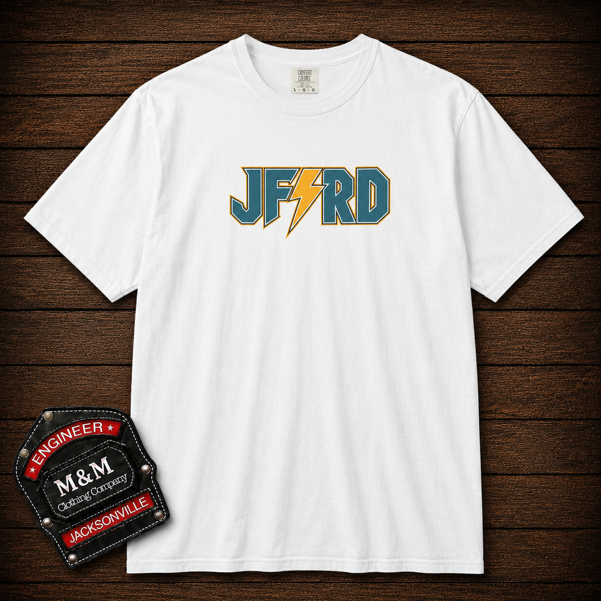 A white short-sleeve crewneck t-shirt with a 'JF&RD' graphic on the chest, featuring teal letters and a yellow lightning bolt. The shirt is displayed flat on a dark wood background, with an 'M&M Engineering Company' badge in the bottom-left corner.