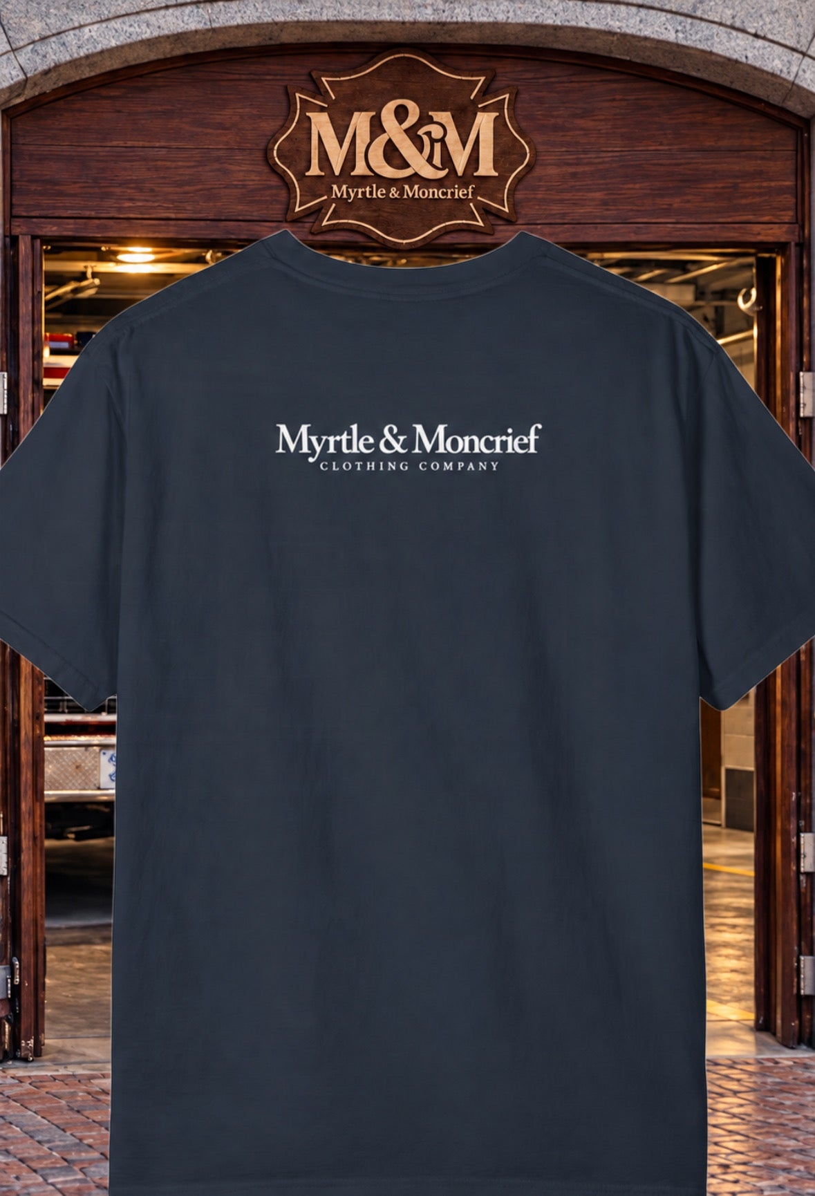 A dark charcoal gray t-shirt with 'Myrtle & Moncrief Clothing Company' printed in white on the back, displayed against a background of a wooden storefront with the same brand logo above.