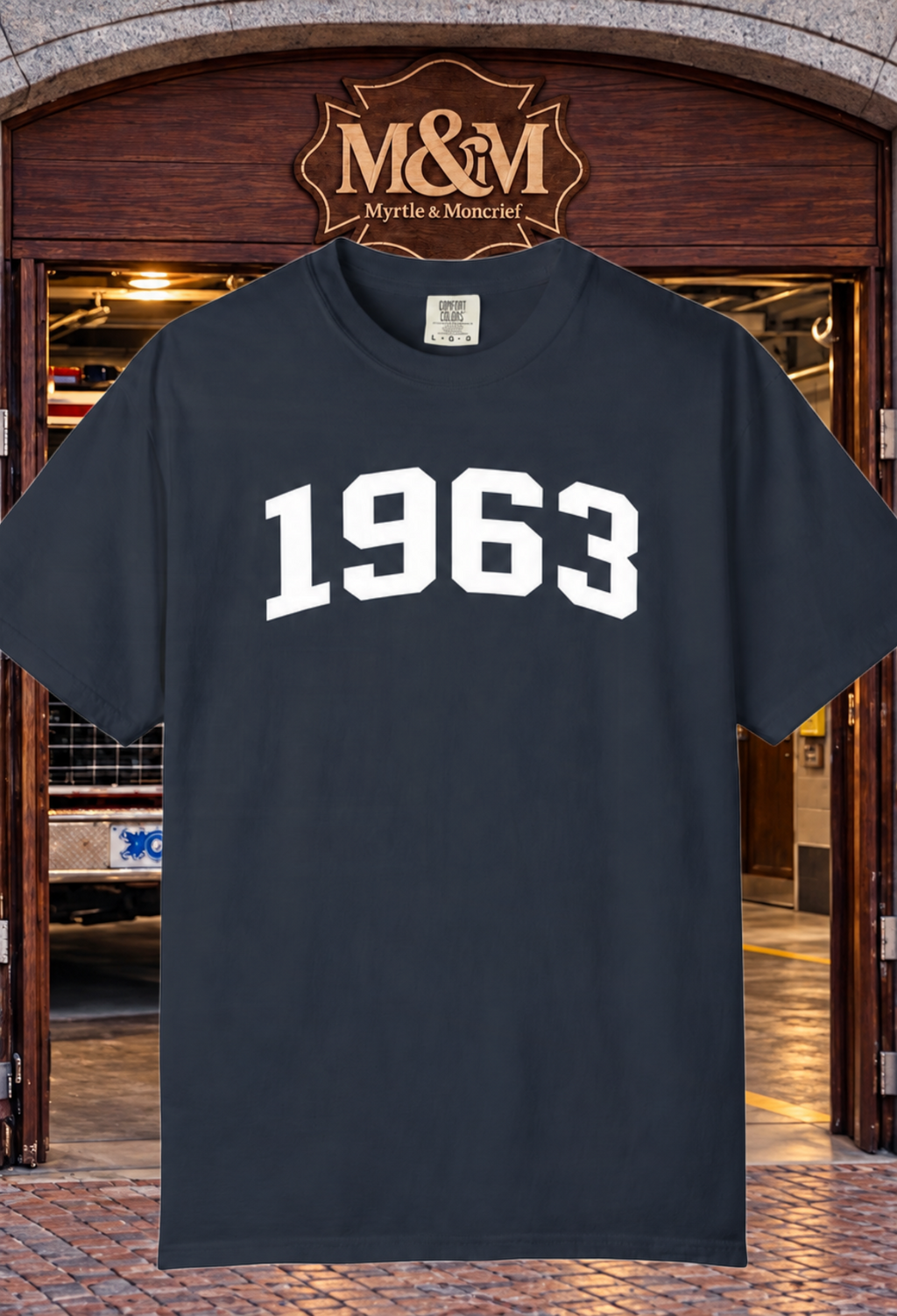 A dark navy blue short-sleeve t-shirt with '1963' printed in white varsity-style block letters across the chest, displayed in front of a wooden door with an 'M&M Myrtle & Moncrief' sign above it.