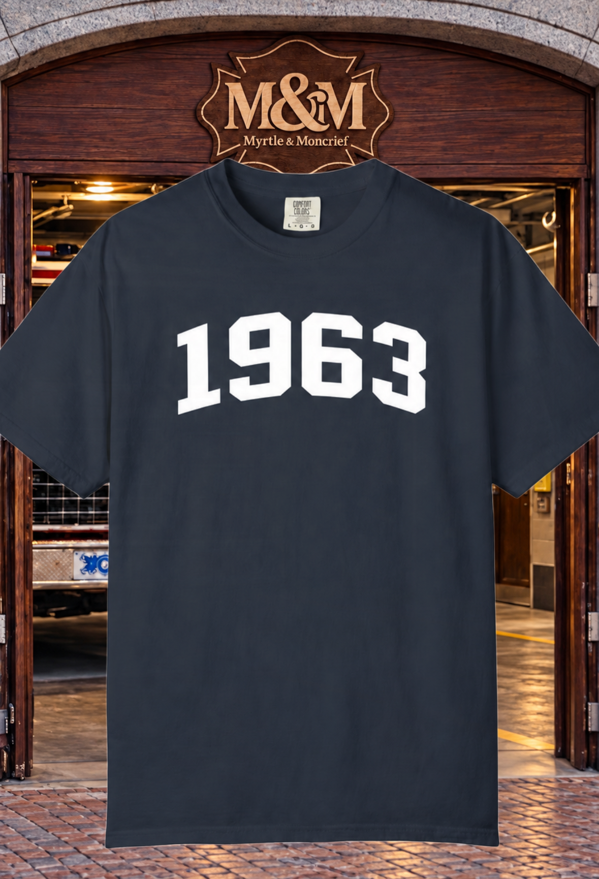 A dark navy blue short-sleeve t-shirt with '1963' printed in white varsity-style block letters across the chest, displayed in front of a wooden door with an 'M&M Myrtle & Moncrief' sign above it.