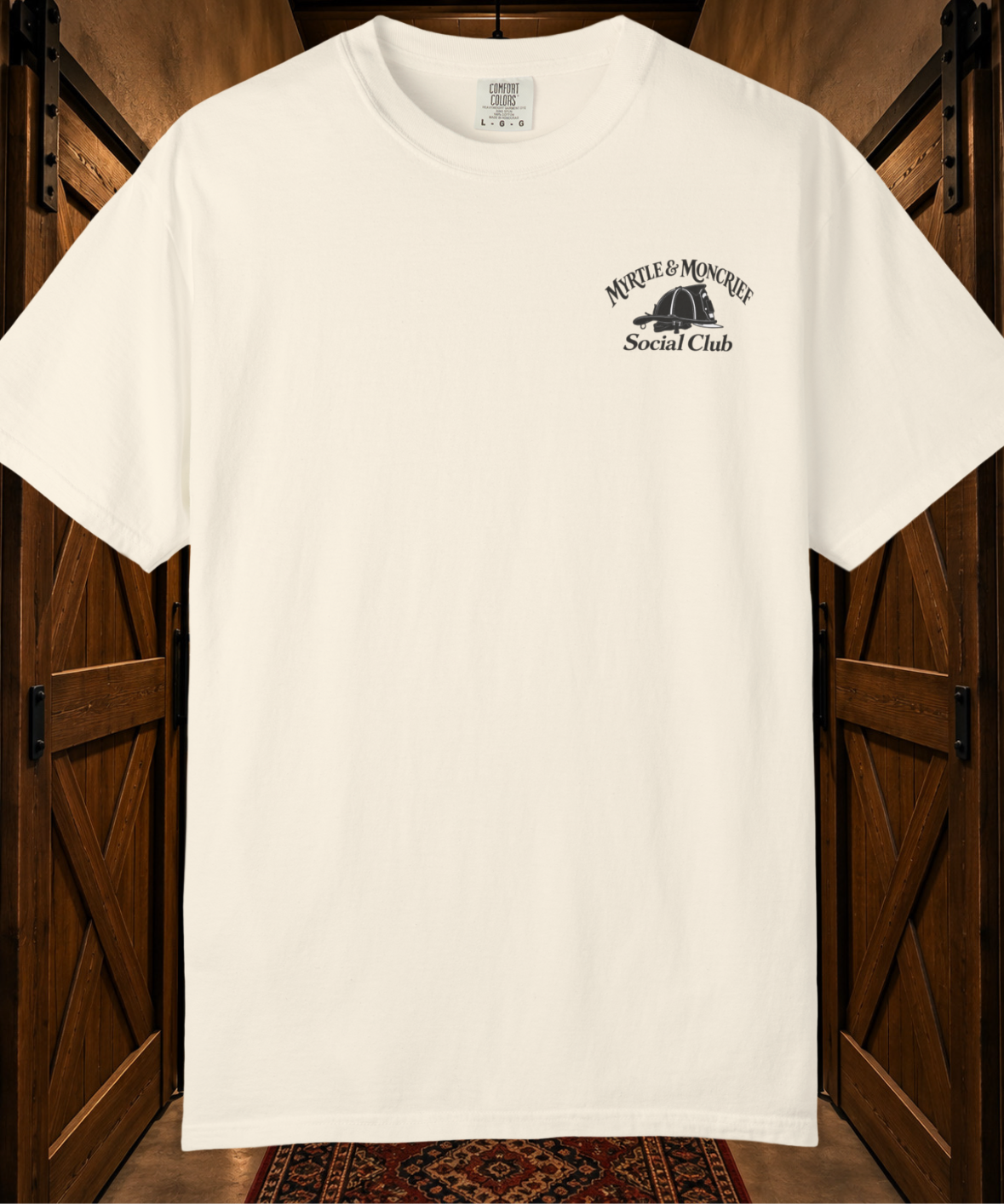 An off-white short-sleeve t-shirt with a black graphic on the left chest reading 'MYRTLE & MONCRIEF Social Club' with a helmet illustration, displayed against a rustic wooden door background.