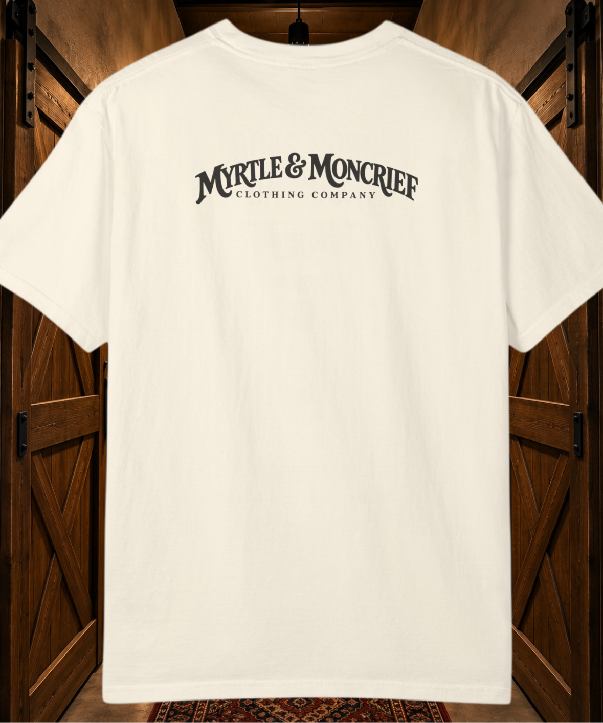 An off-white short-sleeve t-shirt with 'MYRLE & MONCRIEF CLOTHING COMPANY' printed in black across the back, displayed against a rustic dark wood barn door background.