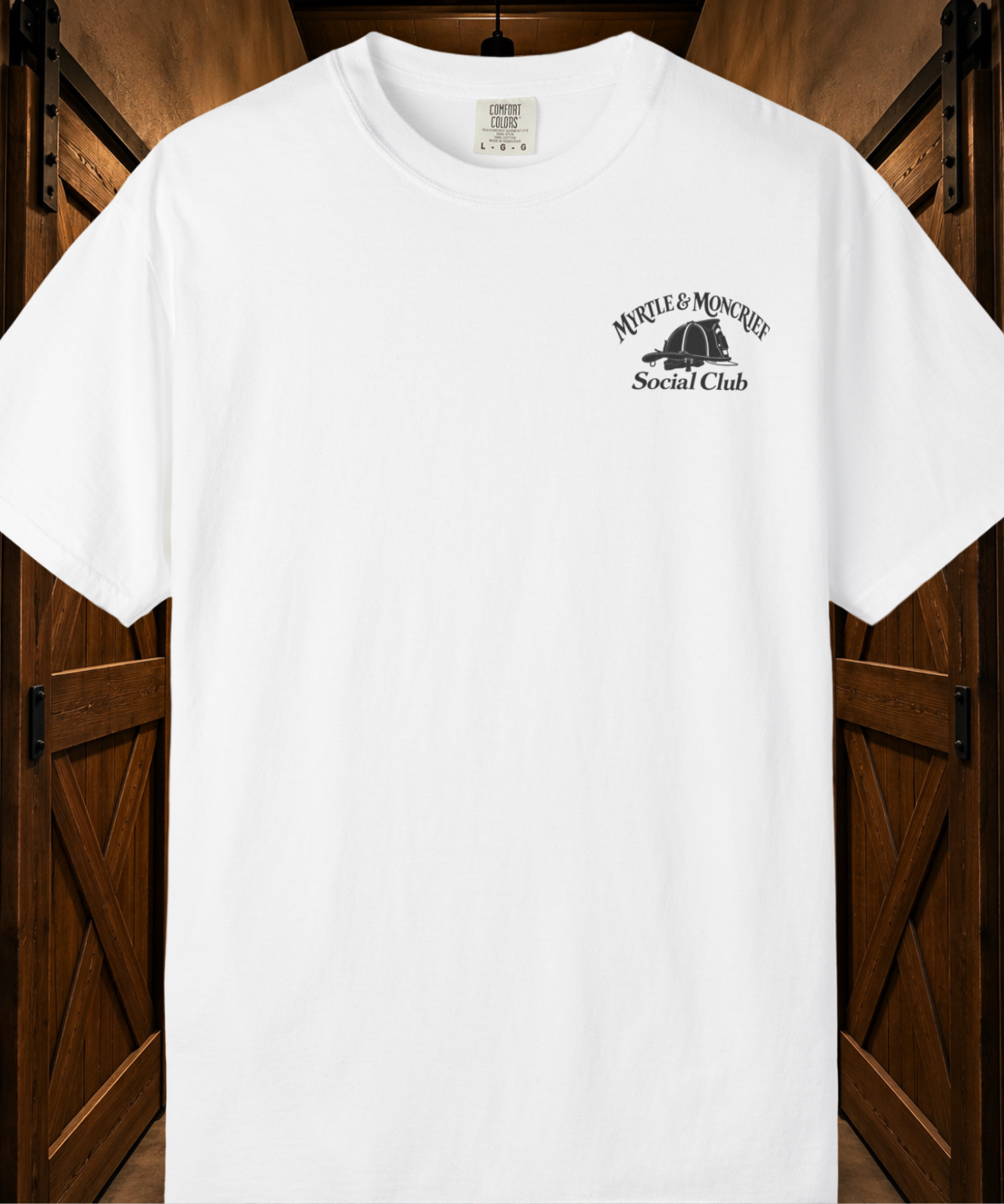 A white short-sleeve crewneck t-shirt with a black graphic on the left chest reading 'MYRTLE & MONCRIEF Social Club' with a fire helmet illustration, displayed against a dark wood barn door background.
