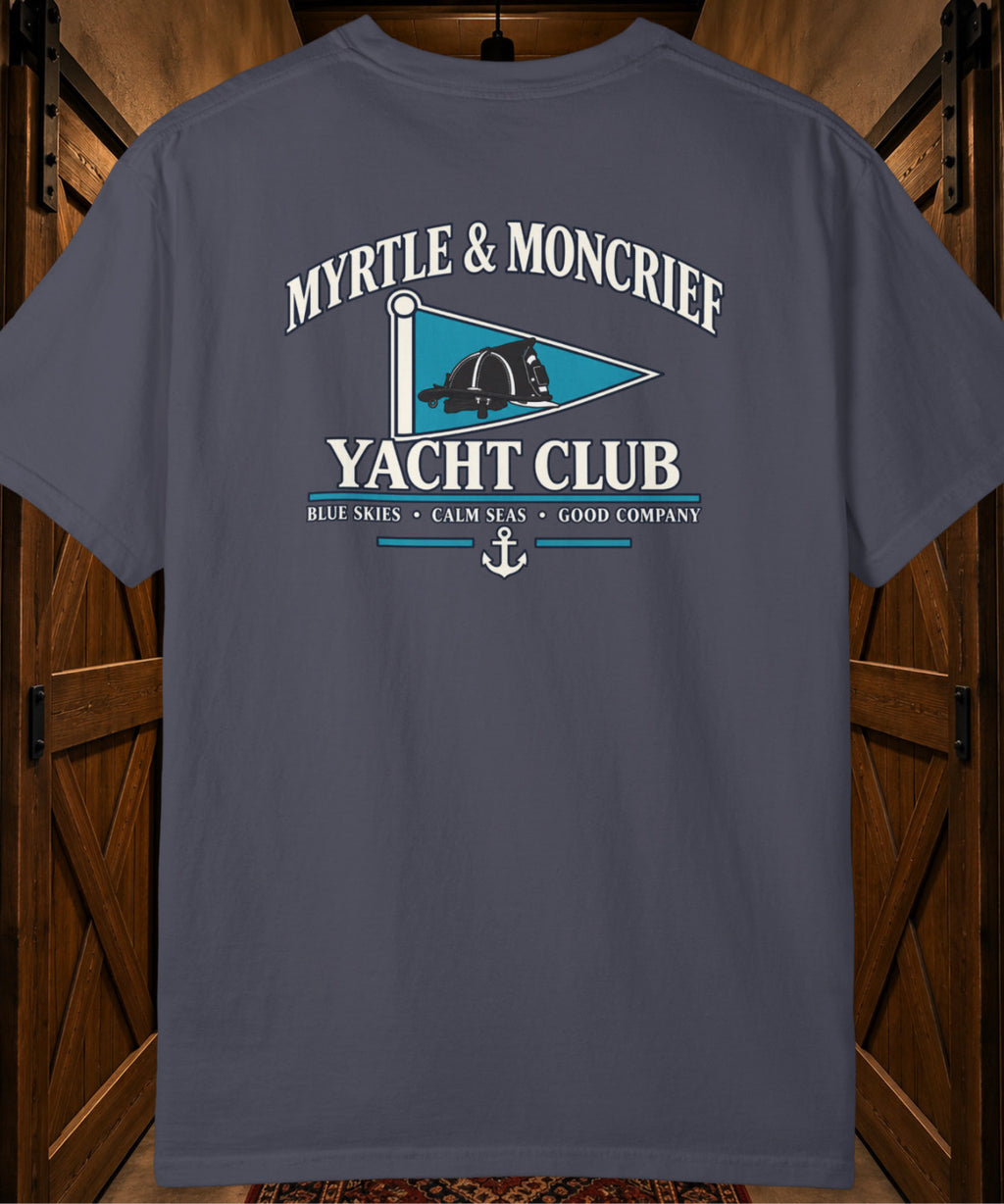 A dark slate blue short-sleeve t-shirt with a vintage-style graphic on the back reading 'MYRTLE & MONCRIEF YACHT CLUB' with a teal flag design, displayed against a dark wood background.
