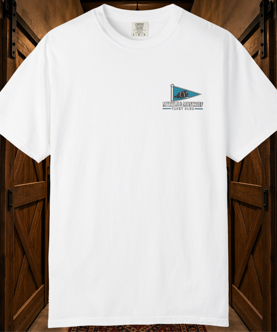 A white short-sleeve crewneck t-shirt with a small chest graphic of a blue and teal flag and the text 'MYRTLE & MONCRIEF YACHT CLUB', displayed against a rustic dark wood background.