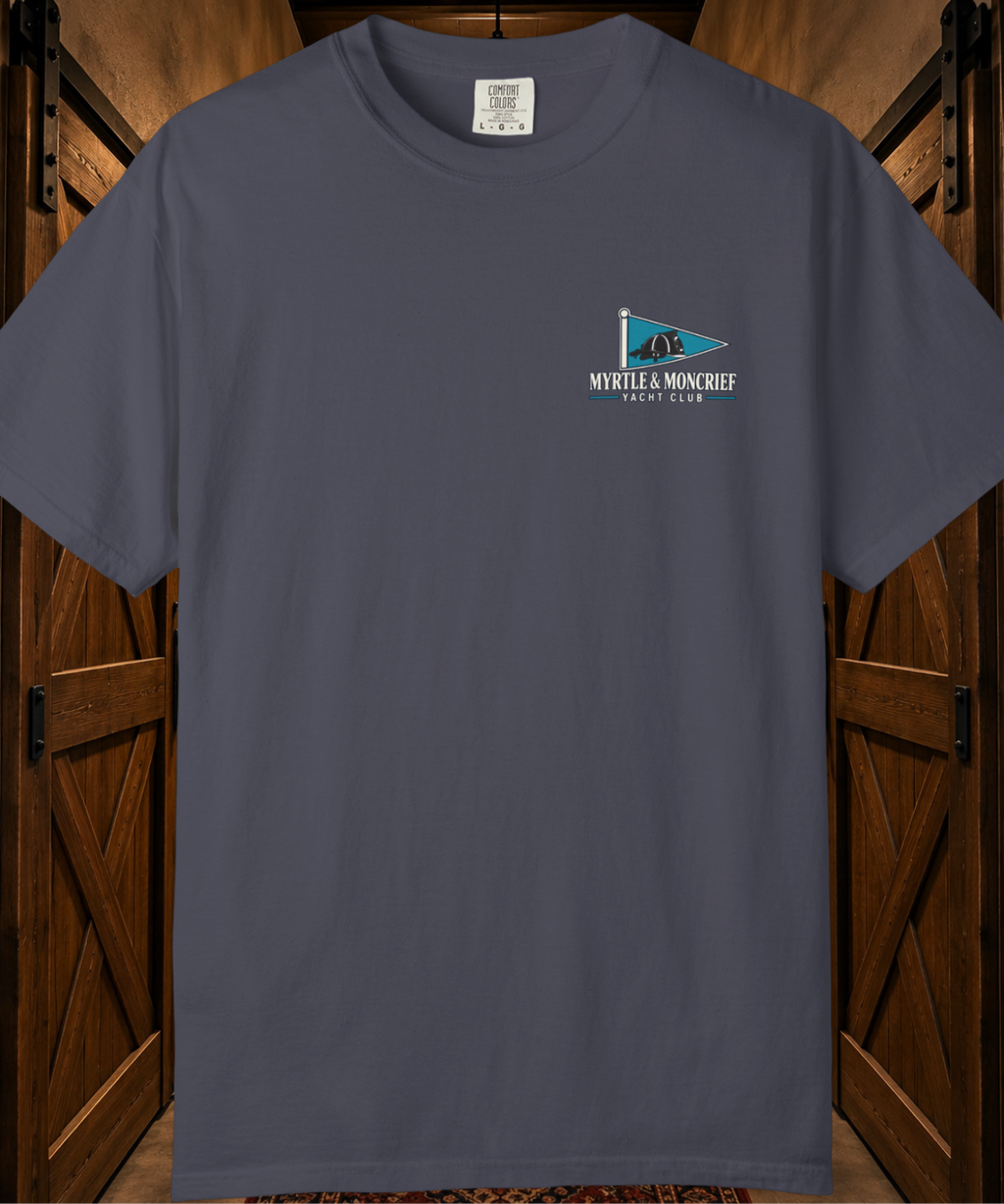A dark slate blue crewneck t-shirt with a small 'MYRTLE & MONCRIEF YACHT CLUB' flag graphic on the left chest, displayed against a rustic wooden barn door background.