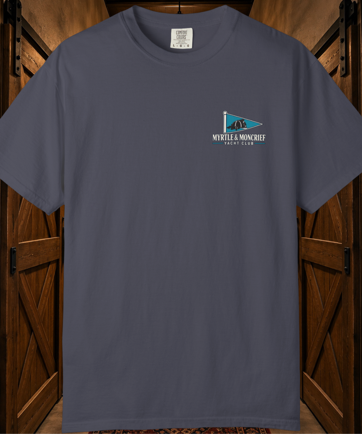 A dark slate blue crewneck t-shirt with a small 'MYRTLE & MONCRIEF YACHT CLUB' flag graphic on the left chest, displayed against a rustic wooden barn door background.