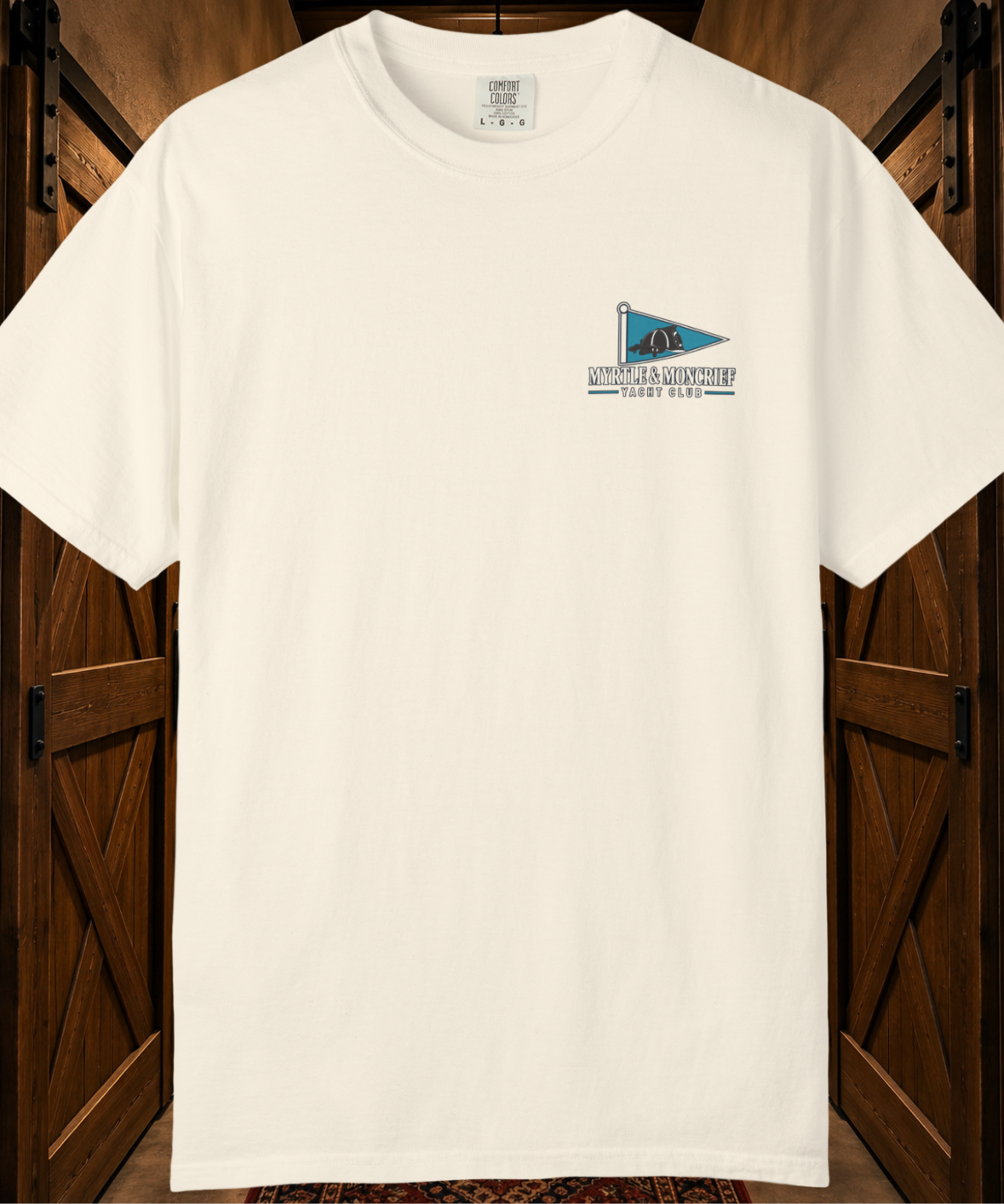 An off-white short-sleeve t-shirt with a small blue and black 'MYRTLE & MONCRIEF YACHT CLUB' logo on the left chest, displayed against a rustic dark wood background.