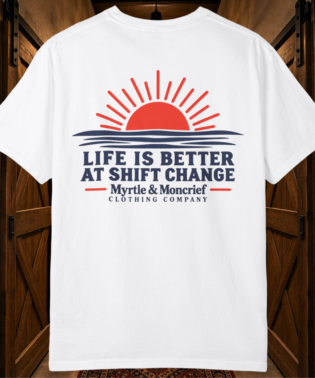 A white short-sleeve t-shirt with a graphic print featuring a red sun over blue waves and the text 'LIFE IS BETTER AT SHIFT CHANGE Myrtle & Moncrief CLOTHING COMPANY', displayed against a dark wood background.