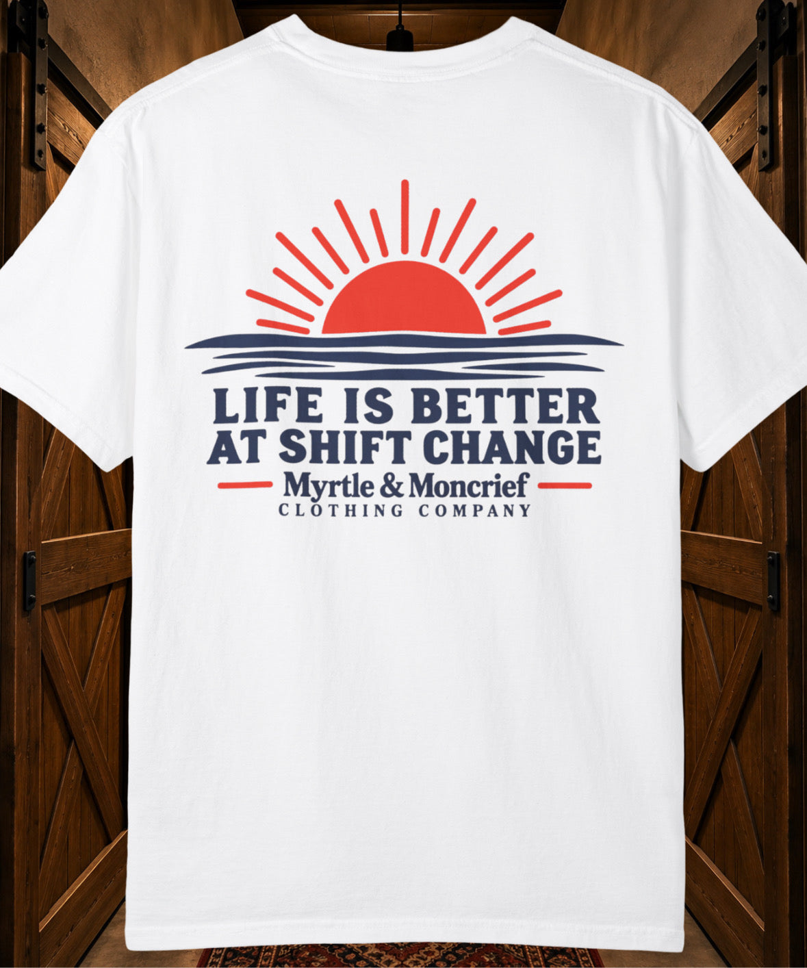 A white short-sleeve t-shirt with a graphic print featuring a red sun over blue waves and the text 'LIFE IS BETTER AT SHIFT CHANGE Myrtle & Moncrief CLOTHING COMPANY', displayed against a dark wood background.