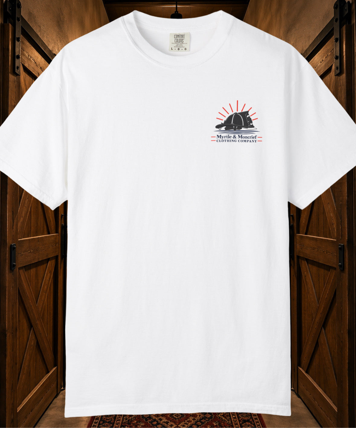 A white short-sleeve crewneck t-shirt hanging against a rustic wooden door background. The shirt features a small graphic on the left chest with a black fire helmet silhouette, red sun rays, and the text 'Myrtle & Moncrief CLOTHING COMPANY'.
