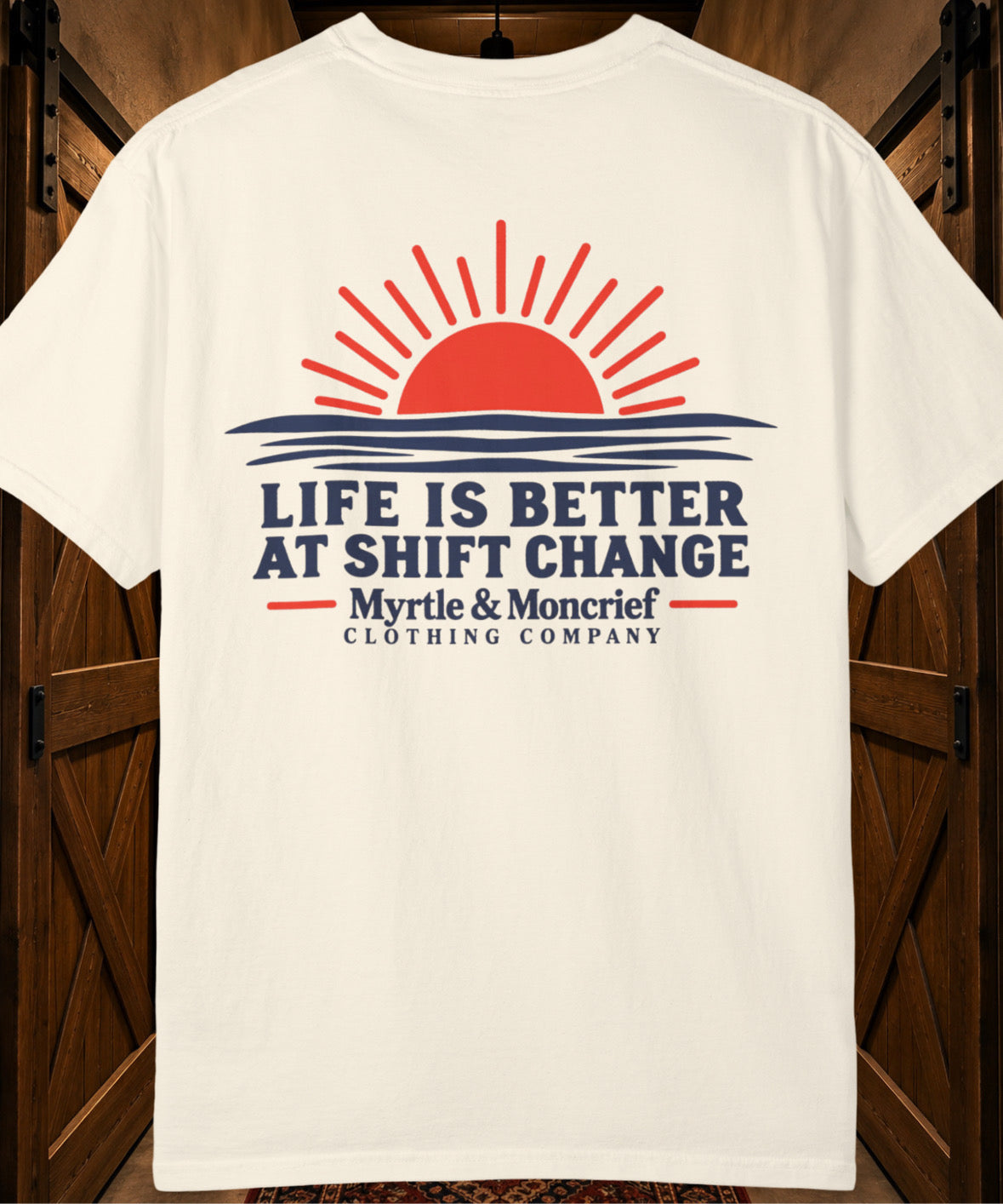 An off-white short-sleeve t-shirt with a graphic print on the back, displayed against a dark wood background. The graphic features a red rising sun over blue waves with the text 'LIFE IS BETTER AT SHIFT CHANGE' and 'Myrtle & Moncrief CLOTHING COMPANY'.