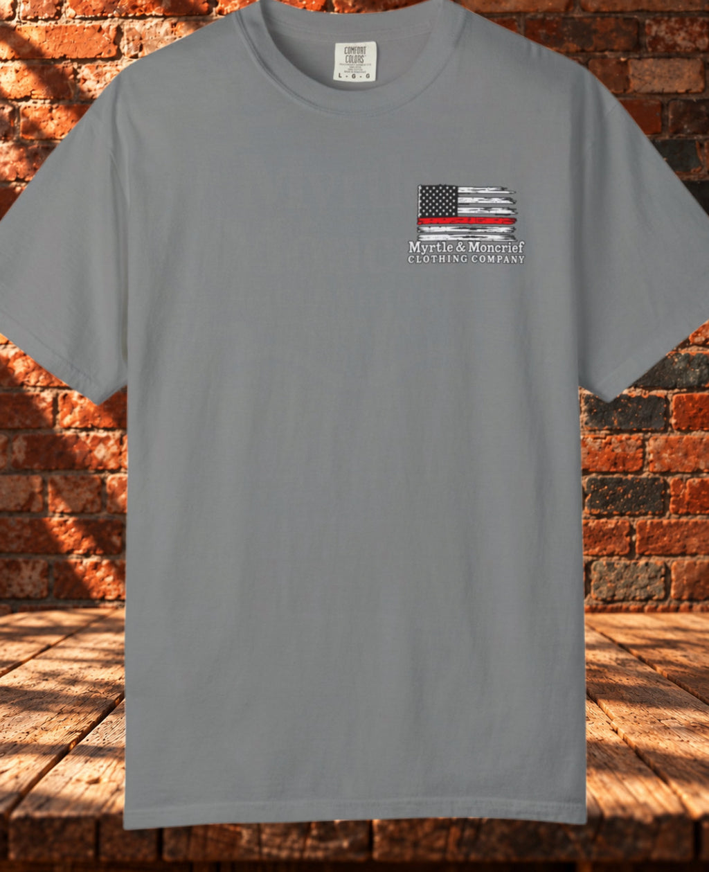 Front view: Gray crewneck T-shirt with a distressed American flag, a single red stripe, and Myrtle & Moncrief branding on the left chest.