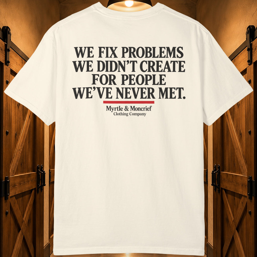 An off-white short-sleeve t-shirt with 'WE FIX PROBLEMS WE DIDN'T CREATE FOR PEOPLE WE'VE NEVER MET.' printed in black text on the back, displayed against a rustic wooden door background.