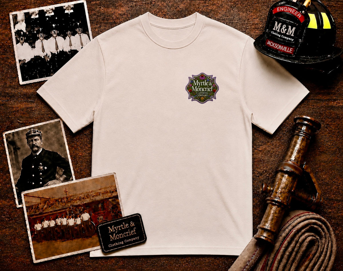 An off-white short-sleeve t-shirt with a Myrtle & Monheit Historic Company emblem on the left chest, styled in a flat-lay on a dark wooden background with vintage firefighter photos, a helmet, a hose reel, and a vintage fire engine.